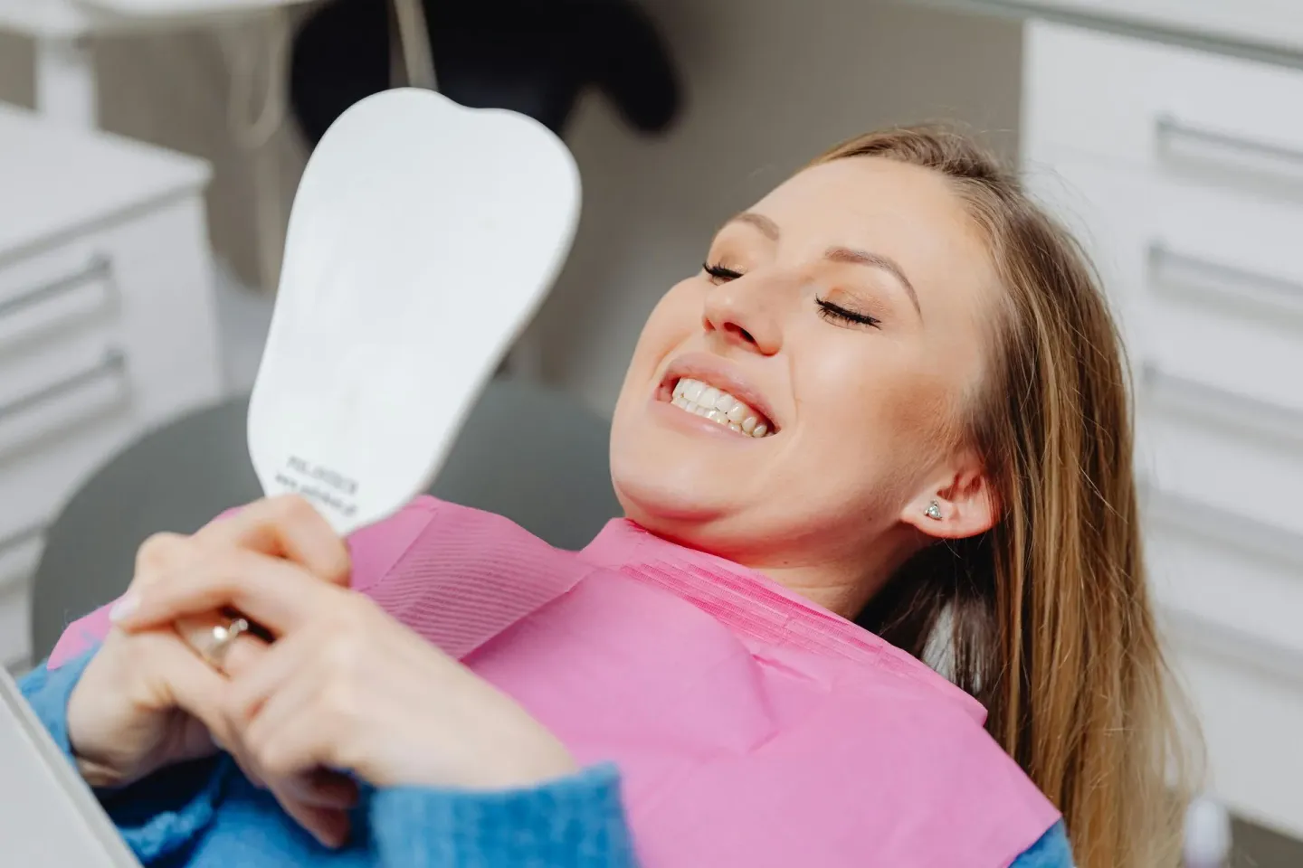A smiling woman in a dental chair looking at her teeth in a hand mirror