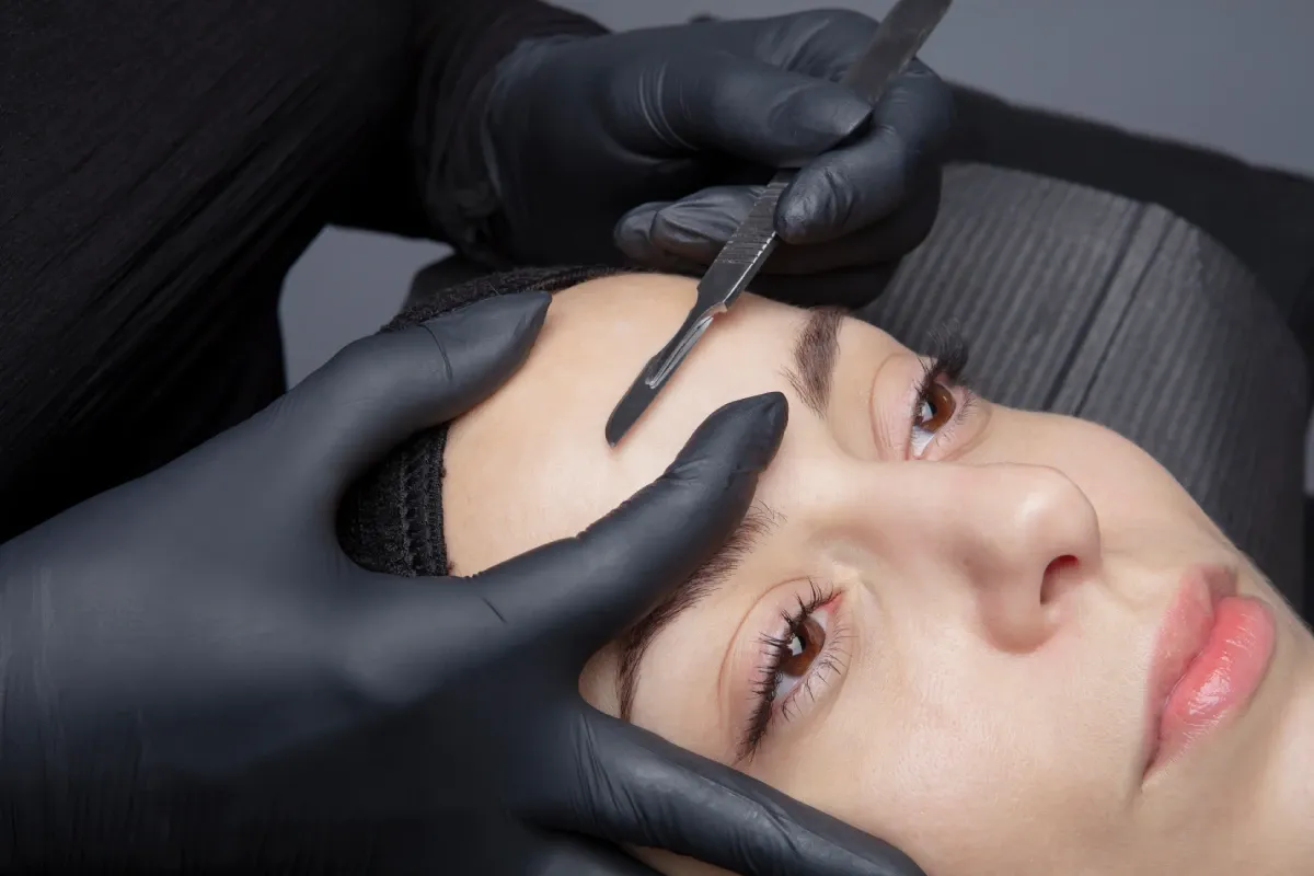Gloved hands holding a small blade to the skin beside a client's brow